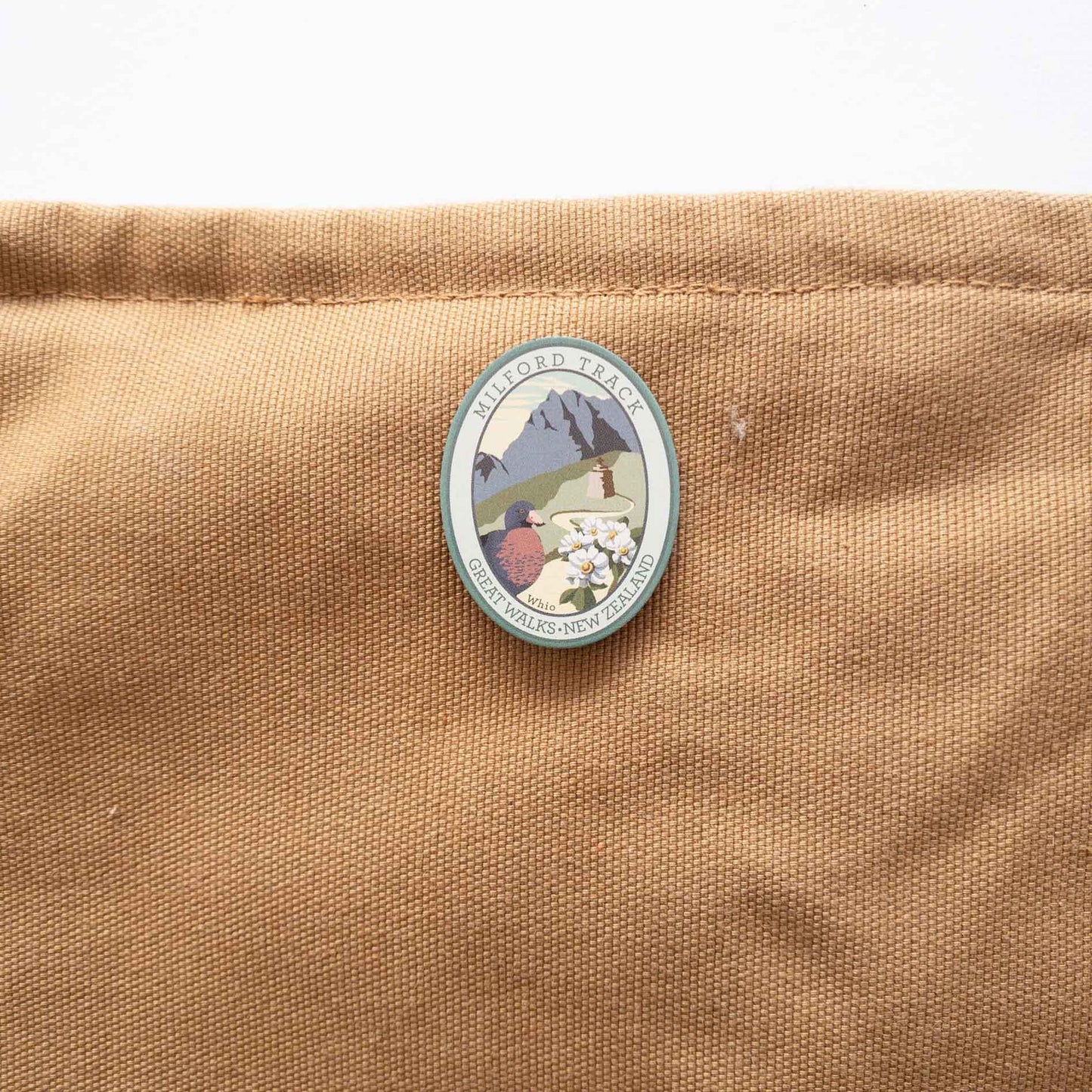 Round Routeburn Track pin, with a mohua/yellowhead bird, purple mountains and blue lake, on a brown canvas bag.