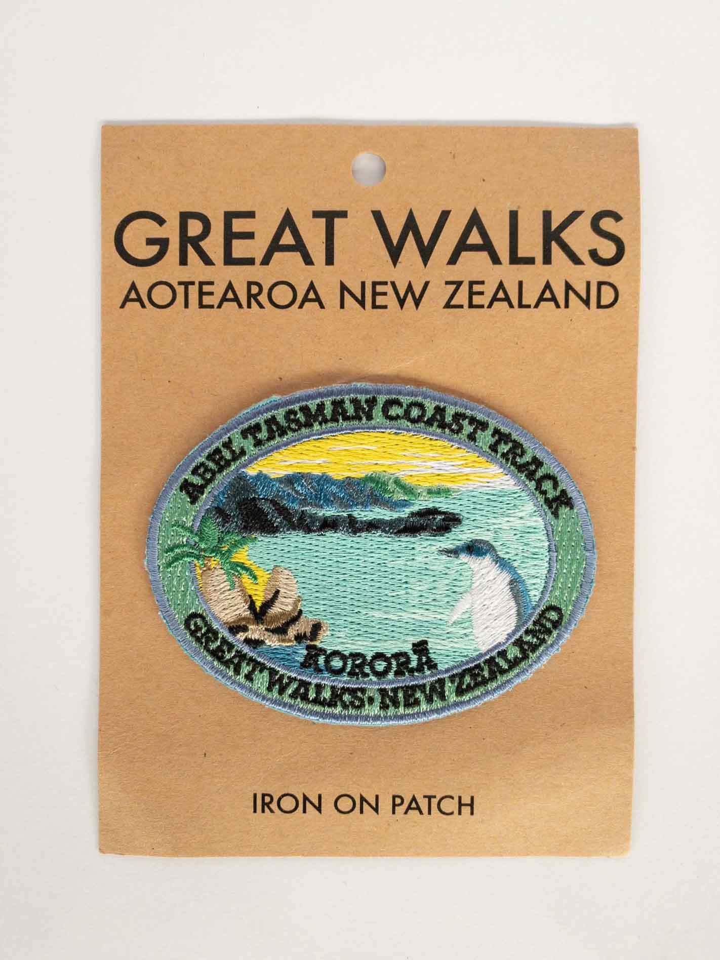 Oval, embroidered Abel Tasman Coast Track patch, with a penguin, blue sea and yellow sky, on brown kraft backing card.