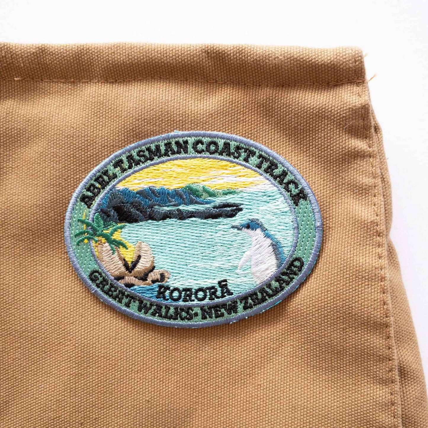 Oval, embroidered Abel Tasman Coast Track patch, with a penguin, blue sea and yellow sky, on a brown canvas bag..