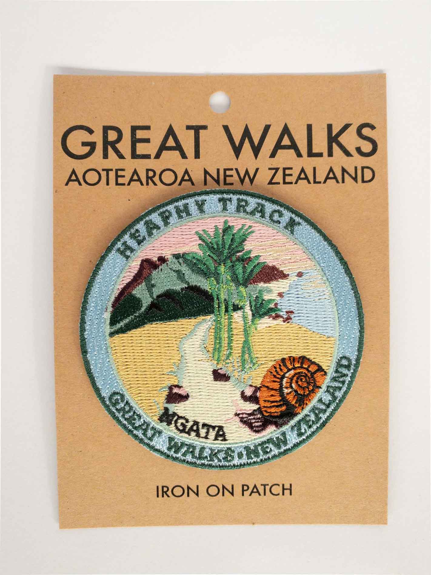 Round, embroidered Heaphy Track patch, with a snail, nikau palms, green hills and pink sky, on brown kraft backing card.