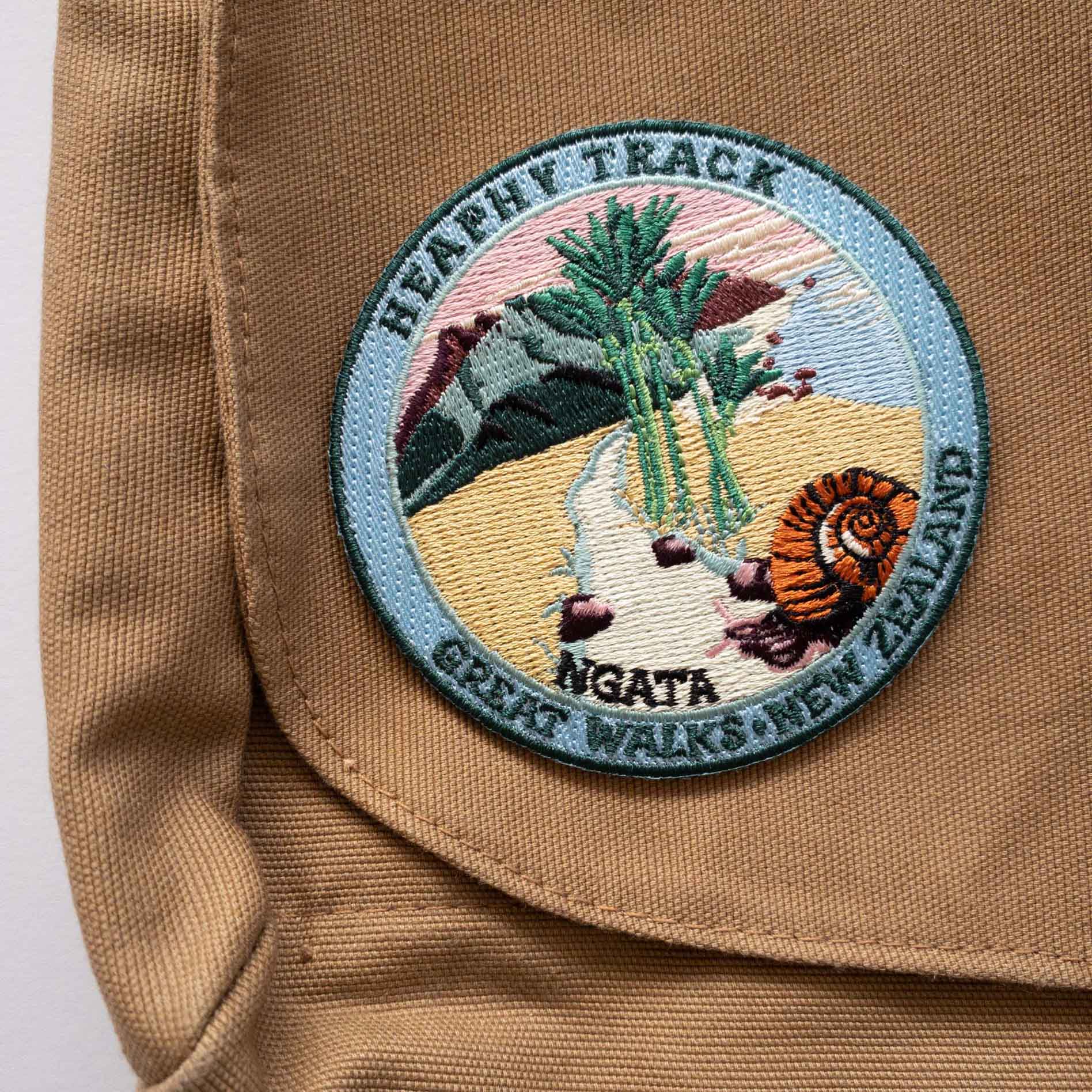 Round, embroidered Heaphy Track patch, with a snail, nikau palms, green hills and pink sky, on a brown canvas bag..