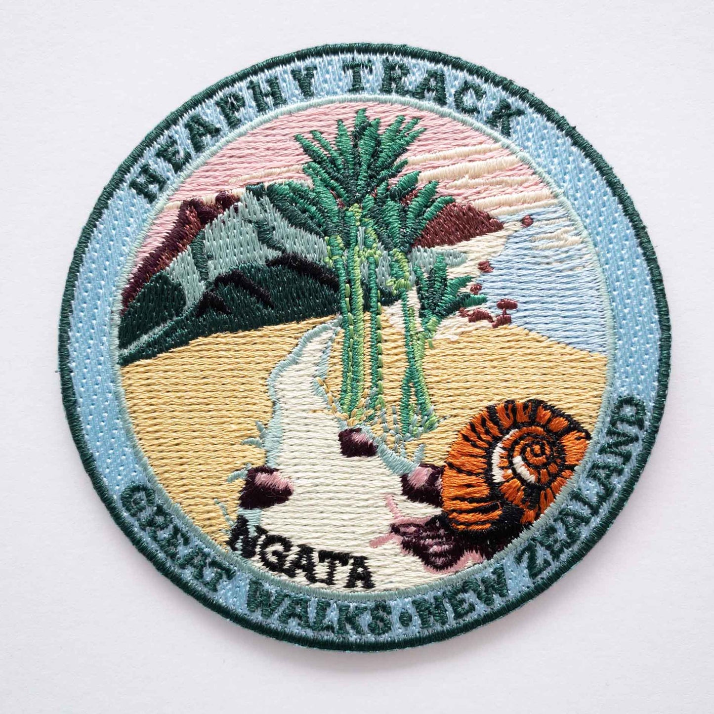 Round, embroidered Heaphy Track patch, with a snail, nikau palms, green hills and pink sky.