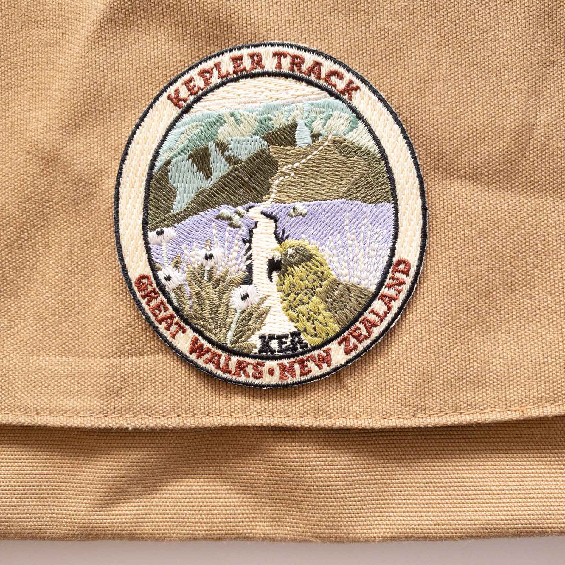 Oval, embroidered Kepler Track patch, with a kea, mountain daisy and alpine ridges, on a brown canvas bag..