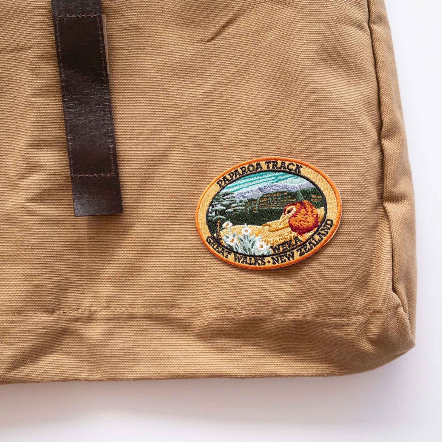 Oval, embroidered Paparoa Track patch, with a weka, green hills, mountain daisy and tussock, on a brown canvas bag.