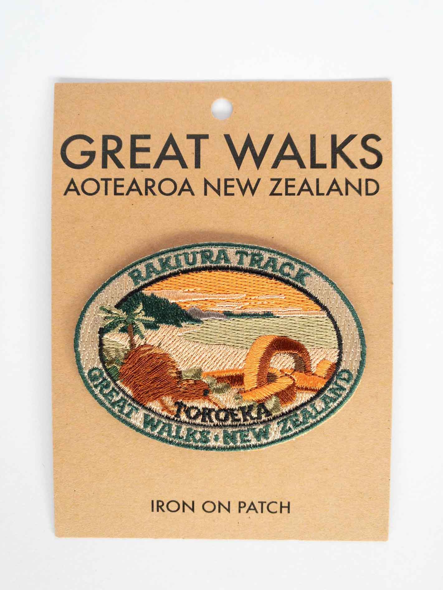 Oval, embroidered Rakiura Track patch, with a brown kiwi, orange sky, and a chain link sculpture, on brown kraft backing card.