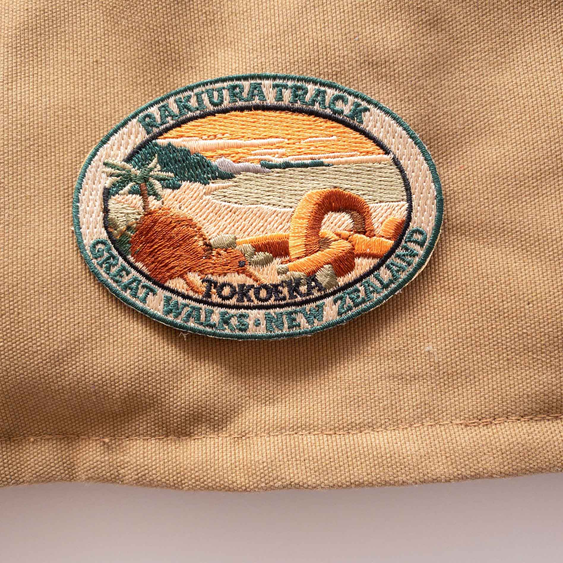 Oval, embroidered Rakiura Track patch, with a brown kiwi, orange sky, and a chain link sculpture, on a brown canvas bag.