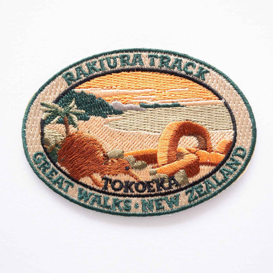 Oval, embroidered Rakiura Track patch, with a brown kiwi, orange sky, and a chain link sculpture.