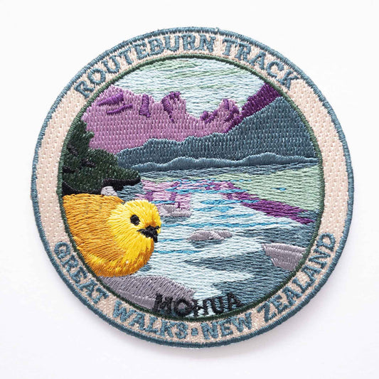 Round, embroidered Routeburn Track patch, with a mohua/yellowhead bird, purple mountains and blue lake.
