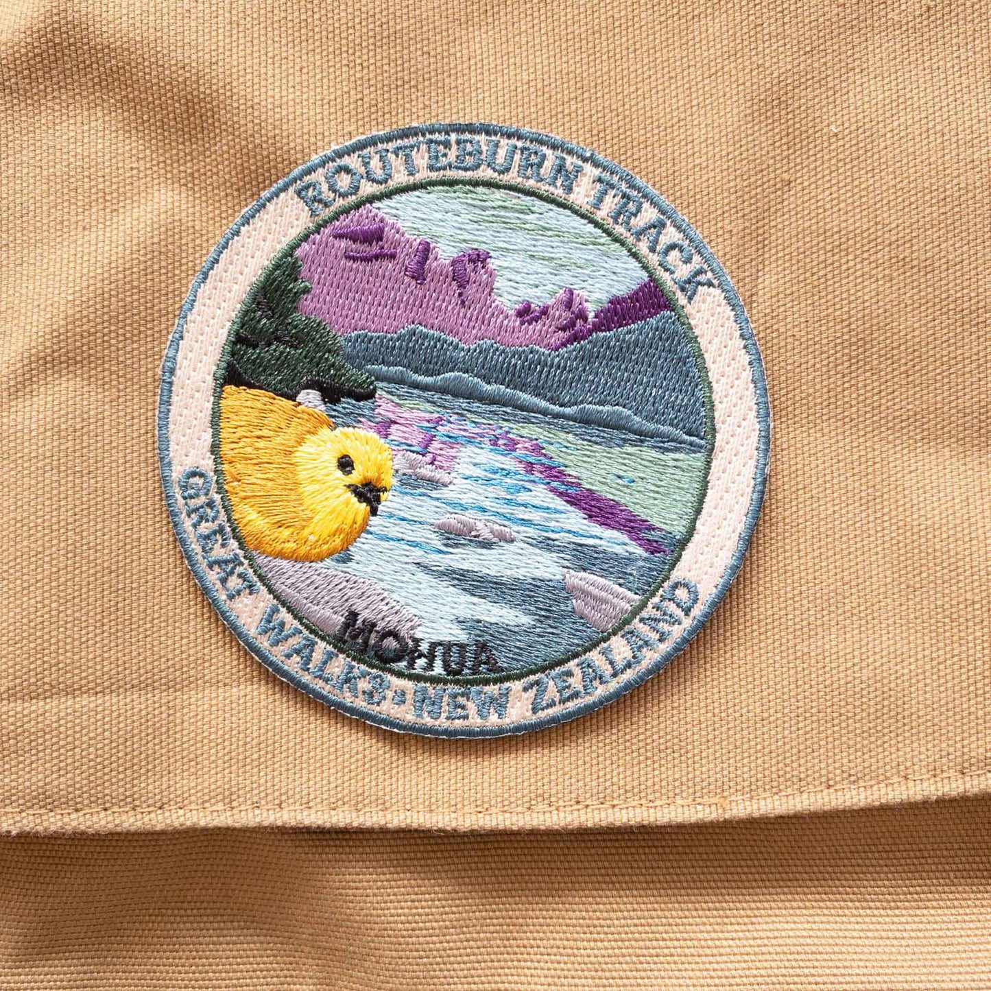 Round, embroidered Routeburn Track patch, with a mohua/yellowhead bird, purple mountains and blue lake, on a brown canvas bag..