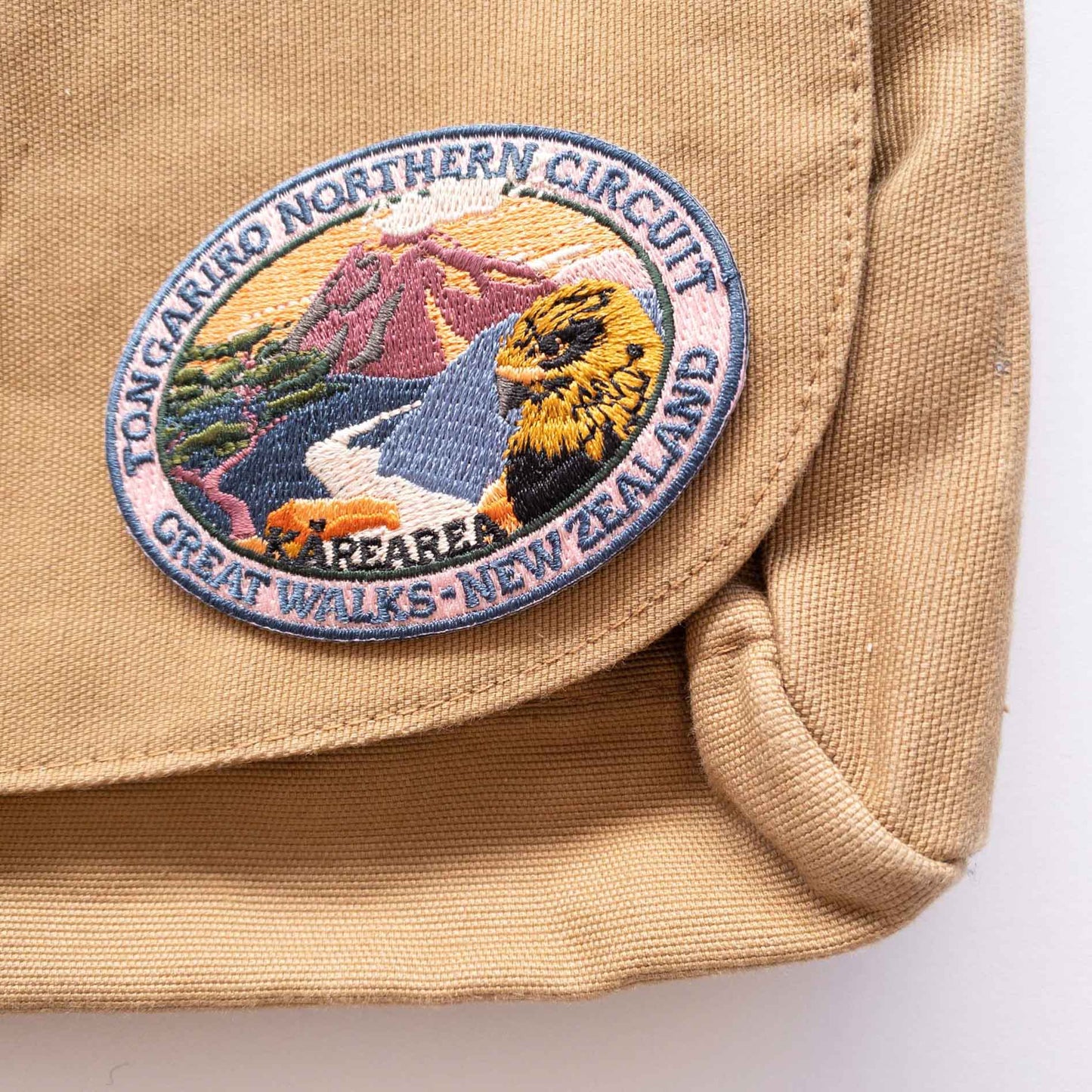 Oval, embroidered Tongaririo Northern Circuit Track patch, with a karearea/falcon, purple active volcano peak and orange sky, on a brown canvas bag.