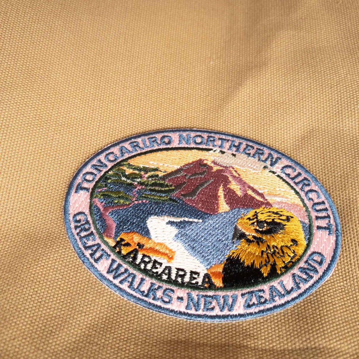 Oval, embroidered Tongaririo Northern Circuit Track patch, with a karearea/falcon, purple active volcano peak and orange sky, on brown kraft backing card.