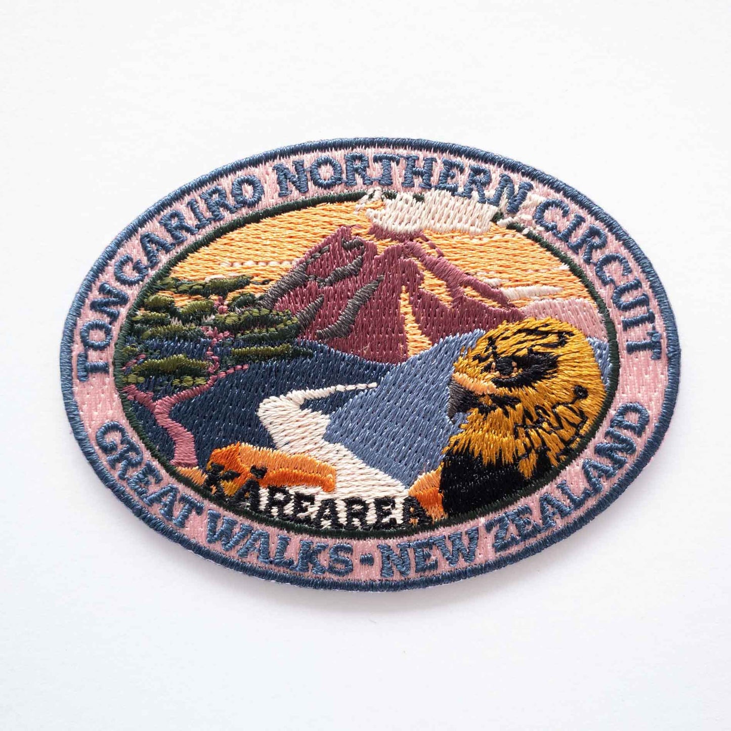 Oval, embroidered Tongaririo Northern Circuit Track patch, with a karearea/falcon, purple active volcano peak and orange sky.