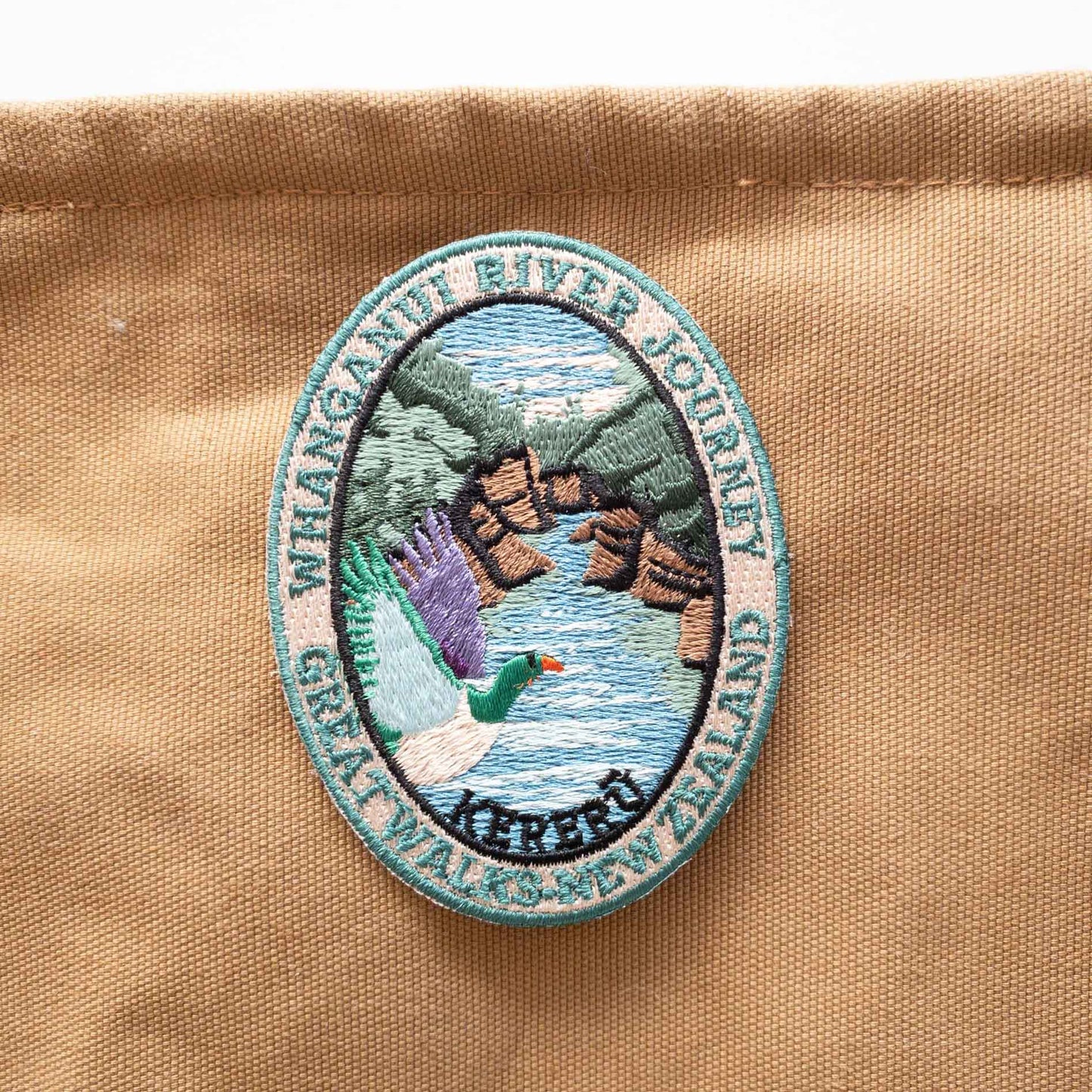 Oval, embroidered Whanganui River Journey Track patch, with a kereru/wood pigeon, green and brown banks and a blue river, on a brown canvas bag.