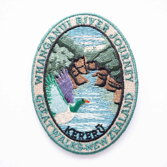 Oval, embroidered Whanganui River Journey Track patch, with a kereru/wood pigeon, green and brown banks and a blue river.