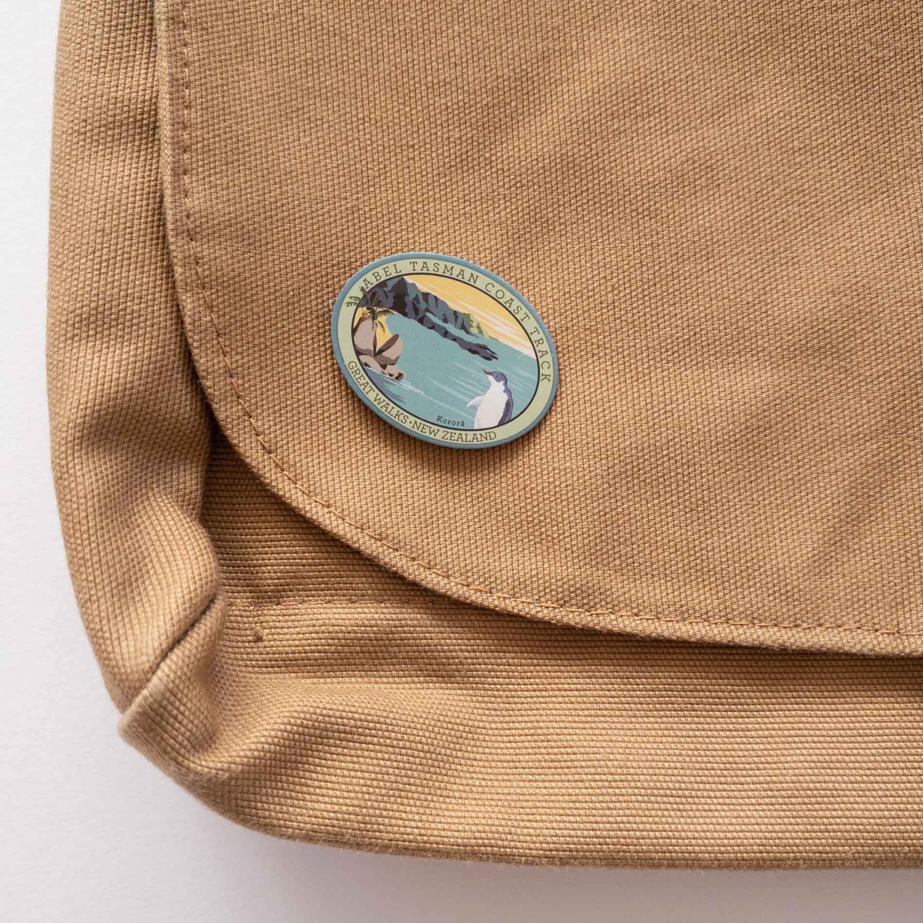 Oval Abel Tasman Coast Track patch, with a little penguin, split apple rock, yellow sky and sandy bays, on a brown canvas bag.