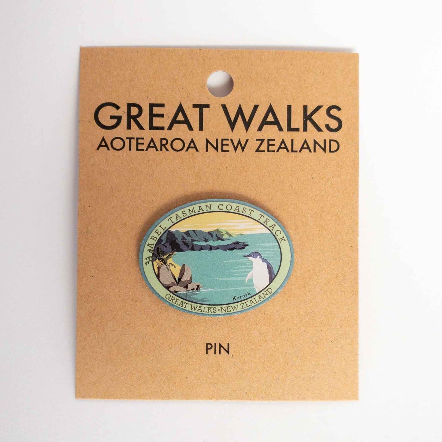 Oval Abel Tasman Coast Track patch, with a little penguin, split apple rock, yellow sky and sandy bays, on brown kraft backing card.