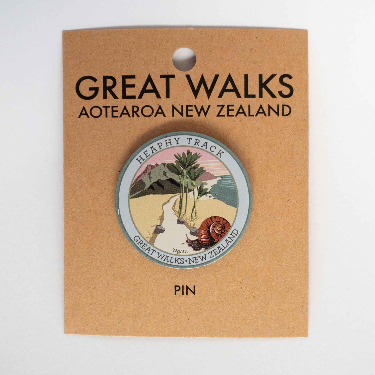 Round Heaphy Track pin, with a snail, nikau palms, green hills and pink sky, on brown kraft backing card.