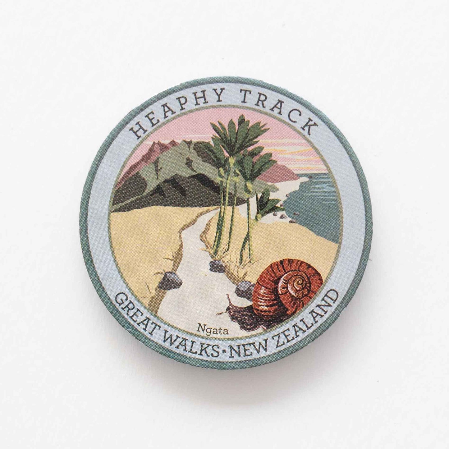 Round Heaphy Track pin, with a snail, nikau palms, green hills and pink sky.