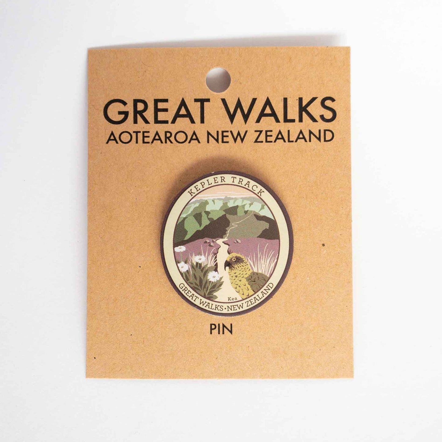 Oval Kepler Track pin, with a kea, mountain daisy and alpine ridges, on brown kraft backing card.