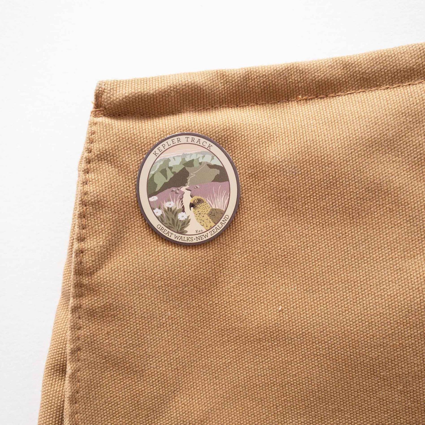 Oval Kepler Track pin, with a kea, mountain daisy and alpine ridges, on a brown canvas bag, on a brown canvas bag.