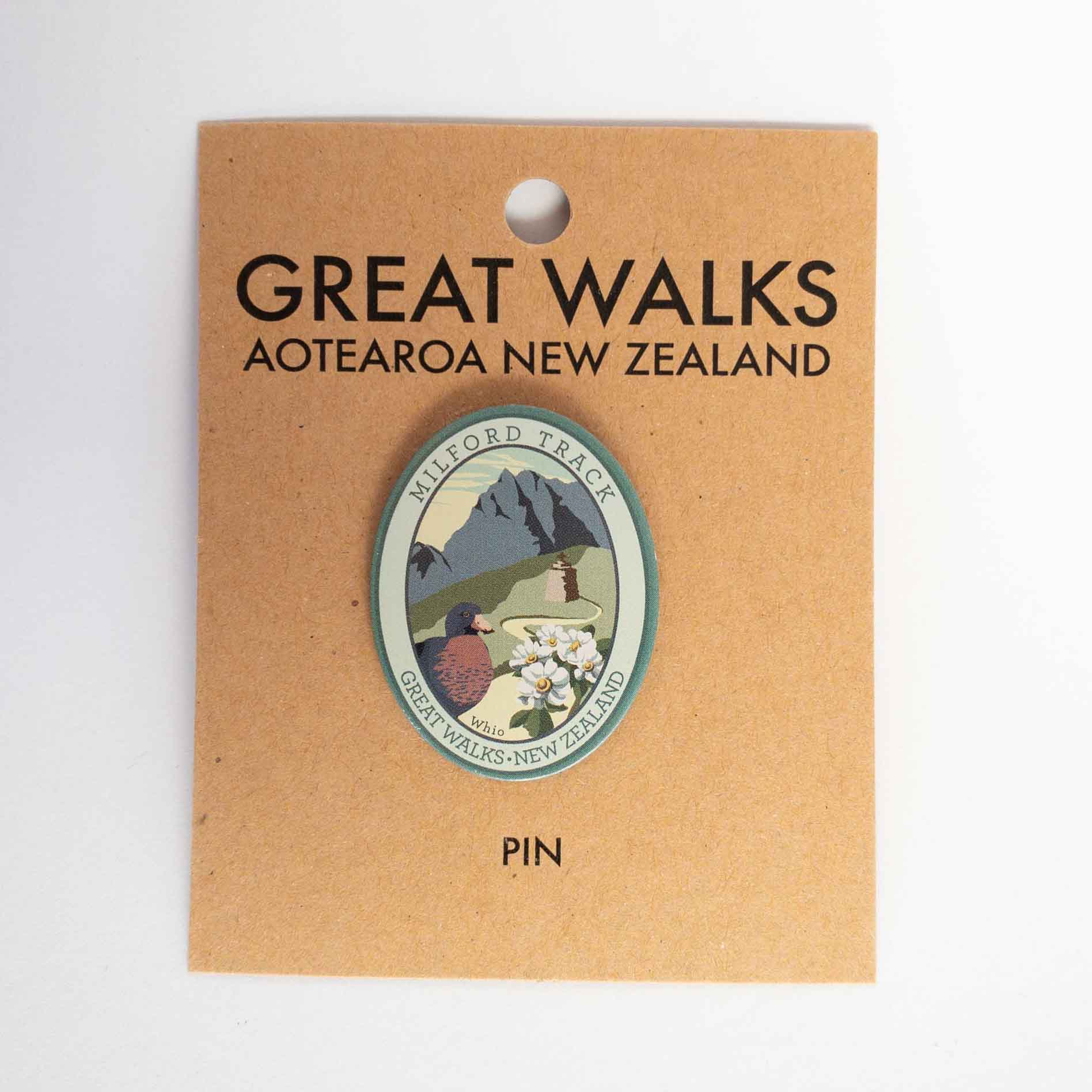 Round Routeburn Track pin, with a mohua/yellowhead bird, purple mountains and blue lake, on brown kraft backing card.