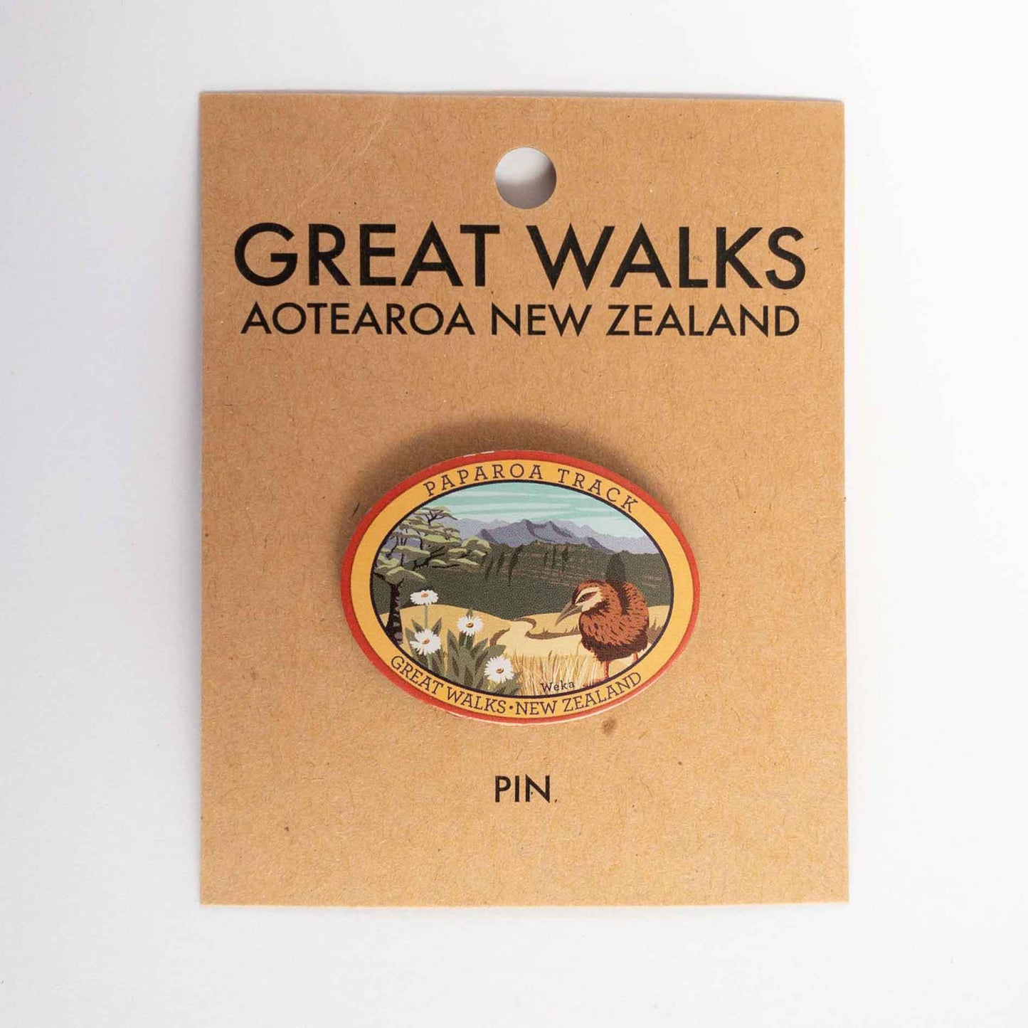 Oval Milford Track pin, with a whio/blue duck, blue peak and mountain buttercup, on brown kraft backing card.