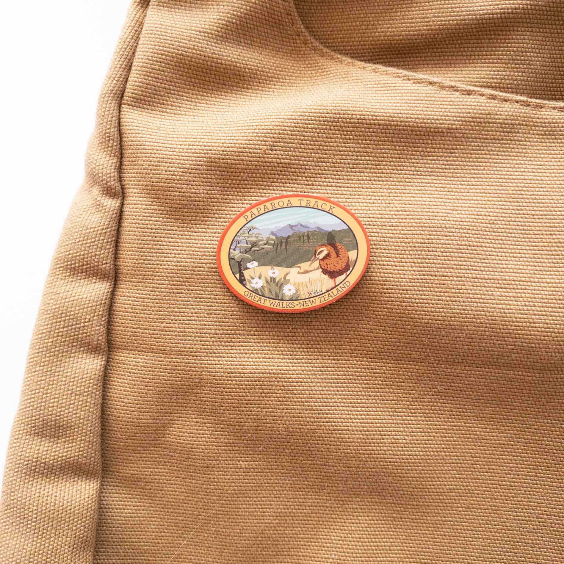 Oval Milford Track pin, with a whio/blue duck, blue peak and mountain buttercup, on a brown canvas bag.
