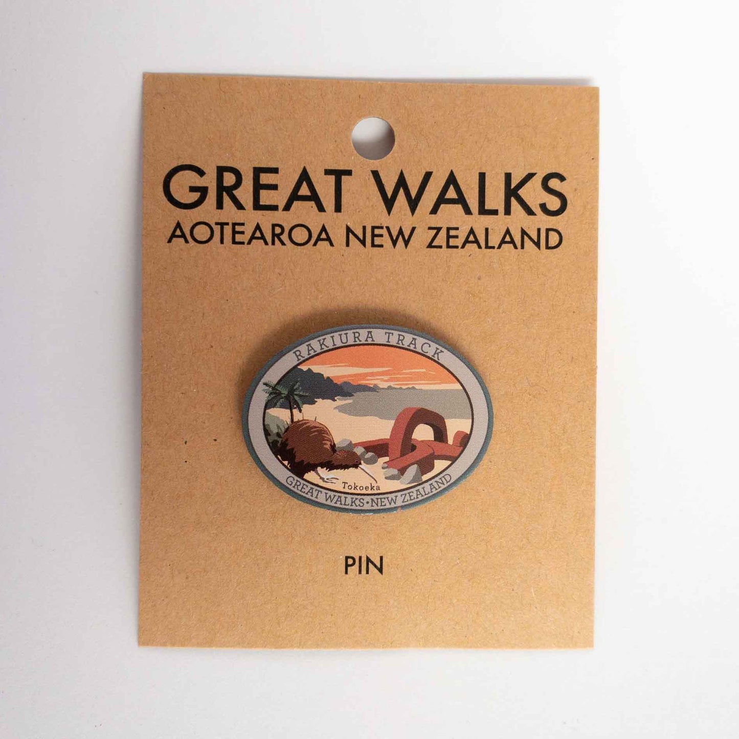 Oval Tongaririo Northern Circuit Track pin, with a karearea/falcon, purple active volcano peak and orange sky, on brown kraft backing card.