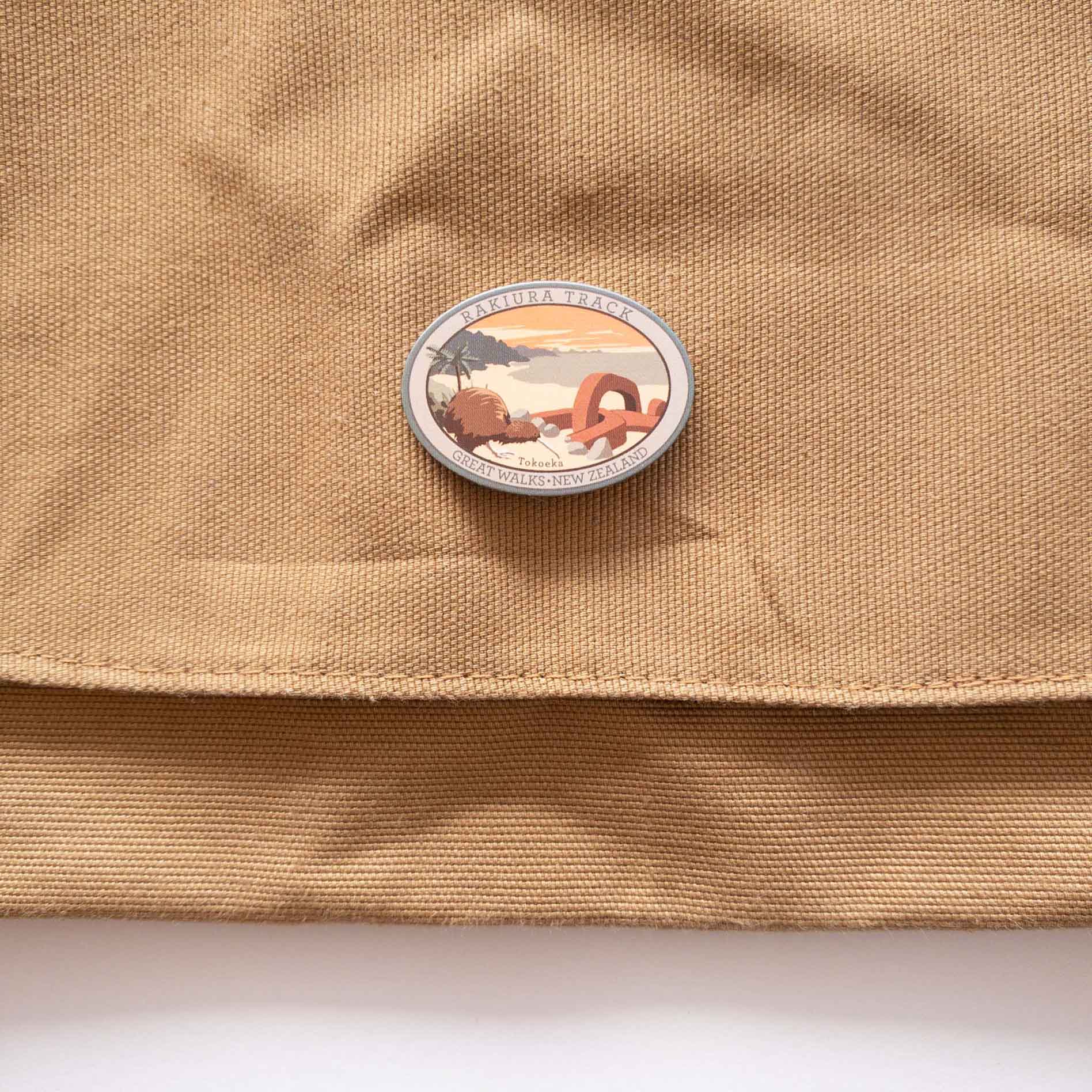 Oval Tongaririo Northern Circuit Track pin, with a karearea/falcon, purple active volcano peak and orange sky, on a brown canvas bag.